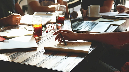 Photo: Personen sitzen an einem Holztisch und arbeiten miteinander. Am Tisch befinden sich Laptops, Bildschirme, Zettel und Stifte 