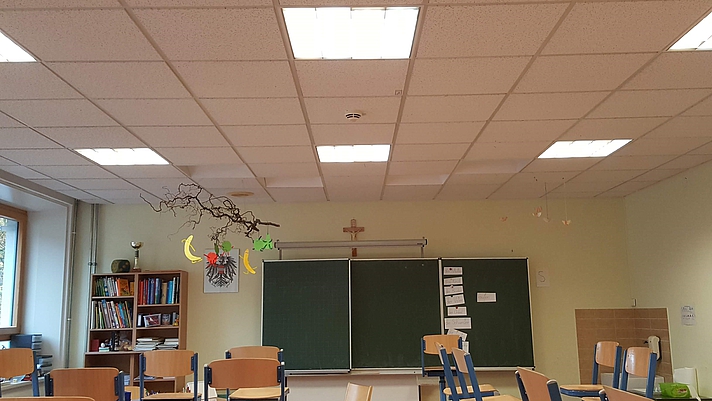 Foto: Volksschulklassenraum mit Stühlen, Tischen und Tafel sowie Deckenbeleuchtungselementen.