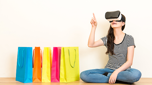 Photo: Frau mit VR Brille zeigt auf was sie kaufen möchte. Seitlich stehen Einkaufstüten
