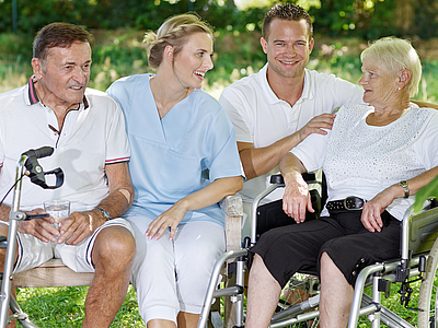 Photo: Eine weibliche und männliche Pflegeperson sitzen im Freien mit einer Patientin und einem Patienten. Die Patientin sitzt in einem Rollstuhl