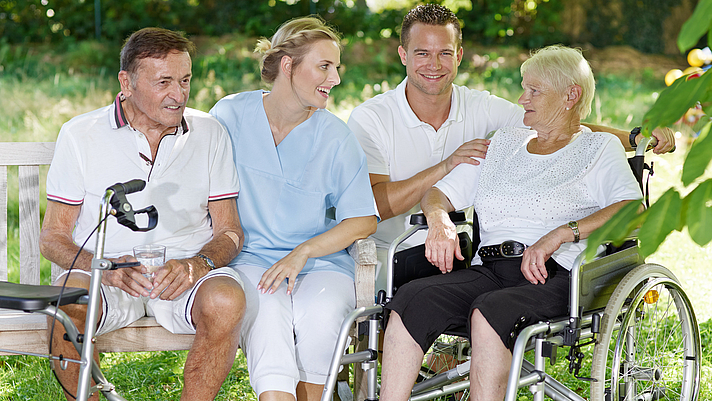 Photo: Eine weibliche und männliche Pflegeperson sitzen im Freien mit einer Patientin und einem Patienten. Die Patientin sitzt in einem Rollstuhl