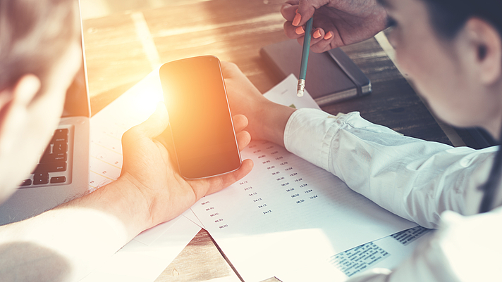 Drei Personen sitzen am Tisch, in der Mitte ein Mobiltelefon