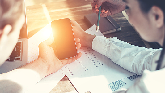 Drei Personen sitzen am Tisch, in der Mitte ein Mobiltelefon