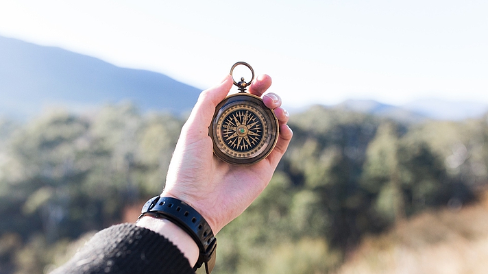 Photo: Hand hält einen Kompass, im Hindergund Natur