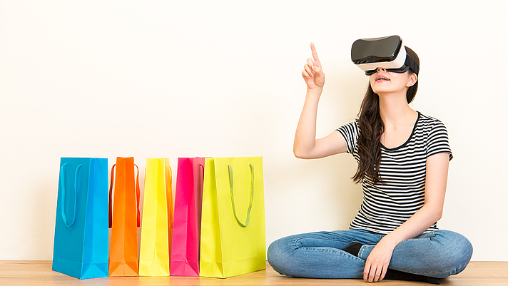 Photo: Frau mit VR Brille zeigt auf was sie kaufen möchte. Seitlich stehen Einkaufstüten