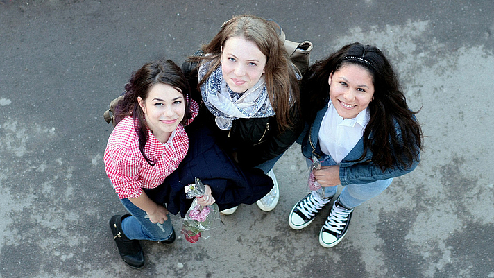 Photo: Drei jungendliche Frauen befinden sich im Freien und blicken lächelnd nach oben in die Kamera 