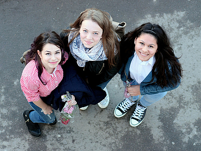 Photo: Drei jungendliche Frauen befinden sich im Freien und blicken lächelnd nach oben in die Kamera 